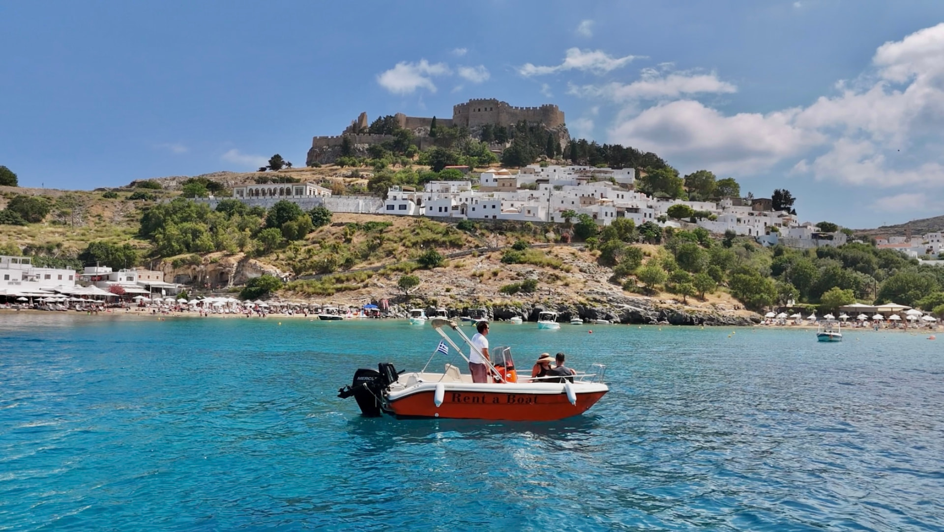 Orange RÉMA rental boat cruising past the Acropolis of Lindos on the Rhodes coastline